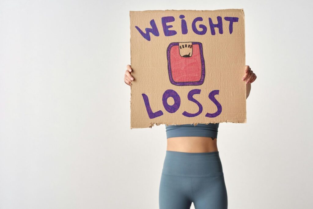 Person in workout gear holding Weight Loss cardboard sign, focusing on health and fitness.