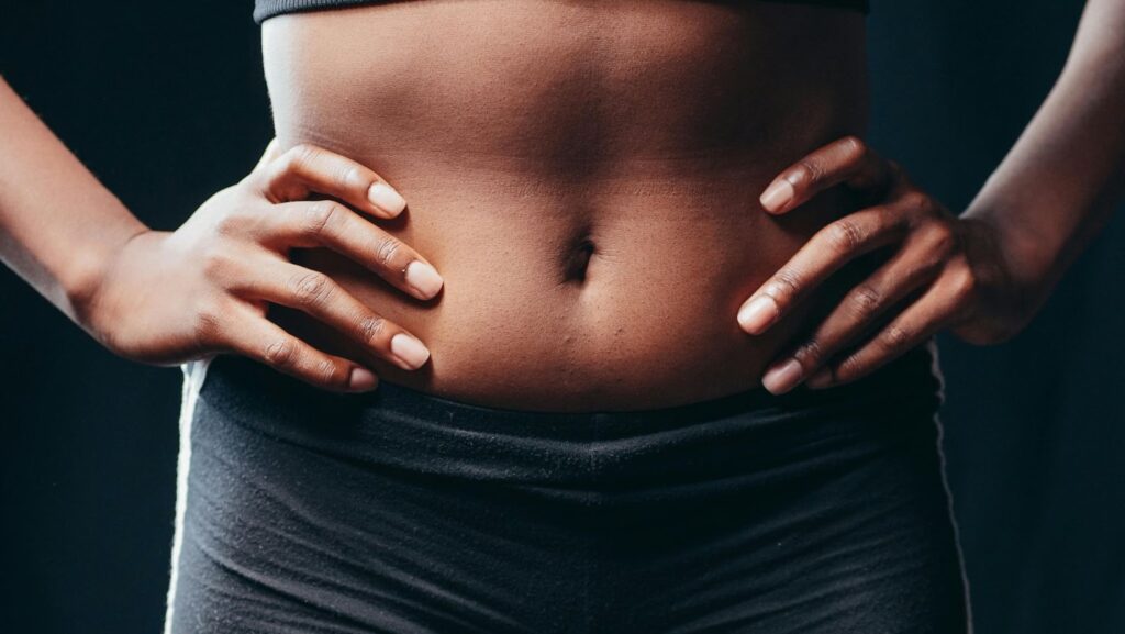 Close-up of a person with hands on hips, showcasing a strong and toned midsection, wearing athletic wear against a dark background.