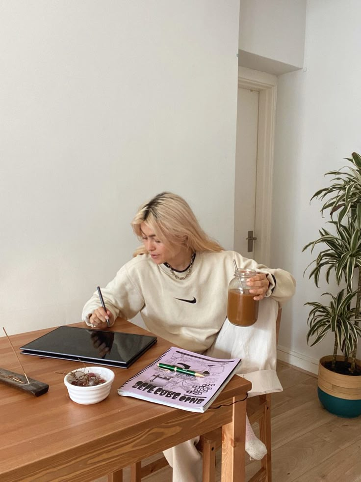 Person drawing on tablet at wooden table with coffee, notebook, and plant. Cozy workspace scene.