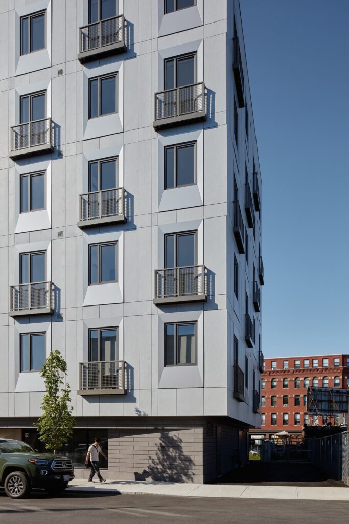 Modern apartment building exterior with balconies, sunny day, urban setting, pedestrian walking by.