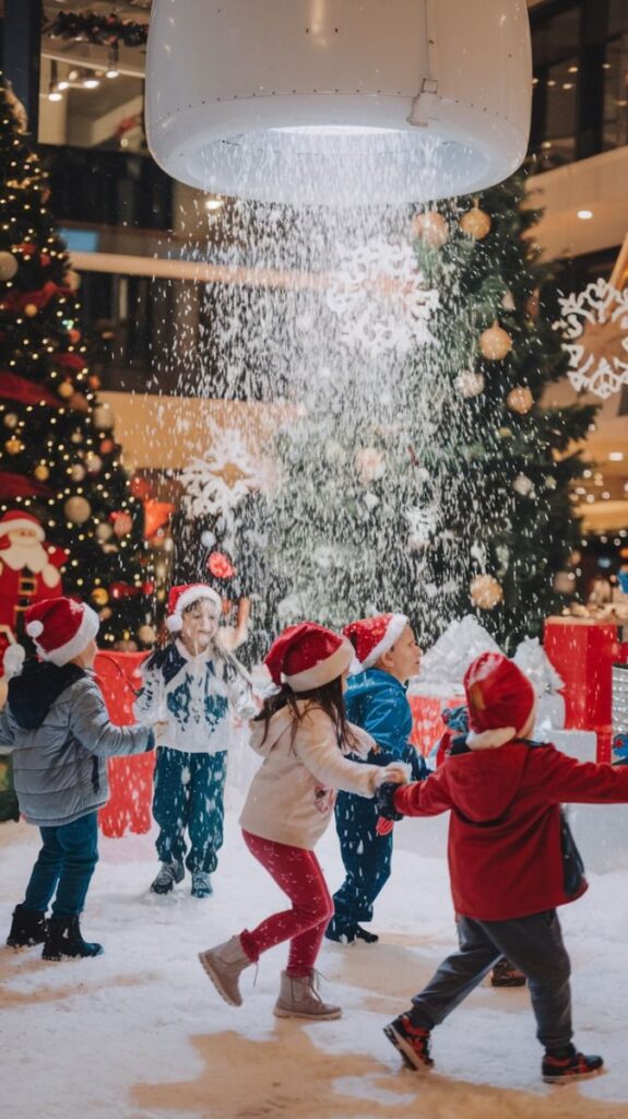 Kids in Santa hats playing in indoor snow, surrounded by Christmas trees and festive decorations.