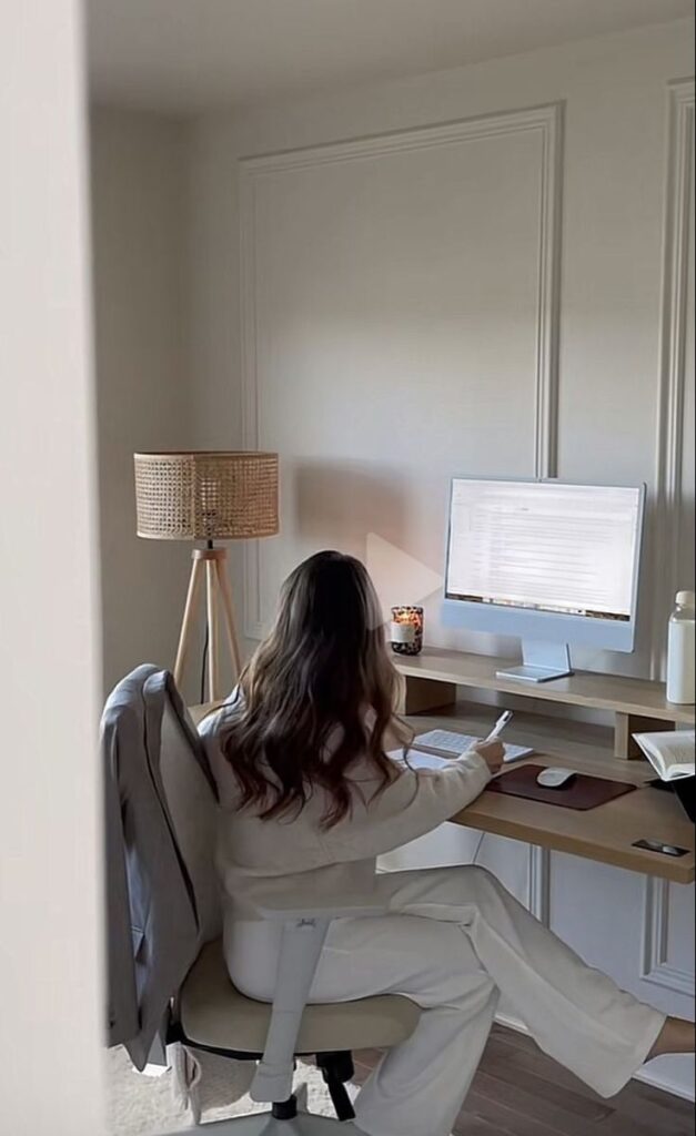 Woman working at a minimalist home office desk with a computer, lamp, and cozy decor elements.