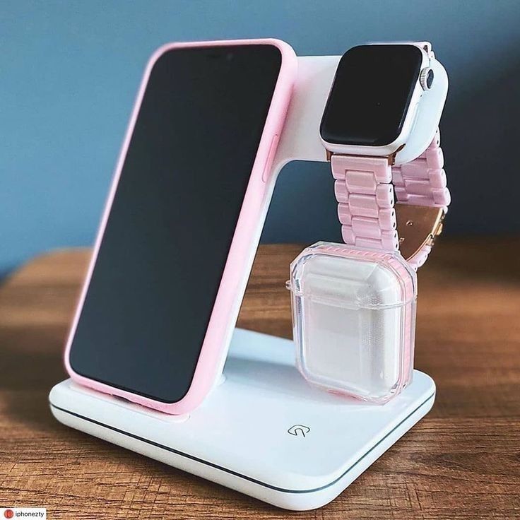 Pink phone, smartwatch, and AirPods displayed on a sleek white charging stand on a wooden surface.