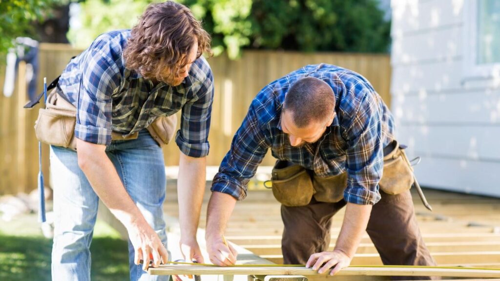 Two construction workers measuring wood outdoors, wearing tool belts and plaid shirts on a sunny day.