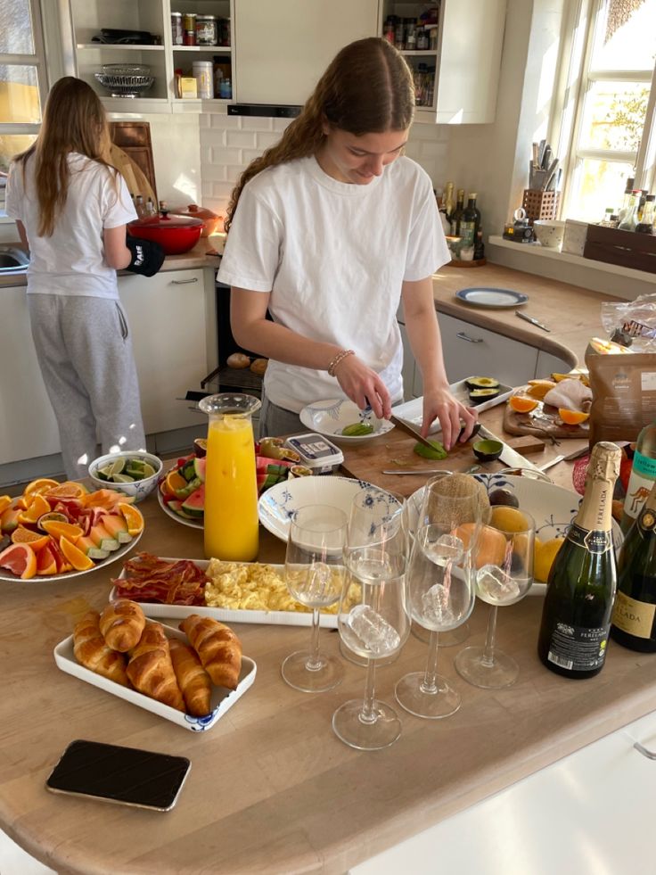 Two people preparing brunch in a cozy kitchen with fresh fruit, pastries, and drinks on the counter.