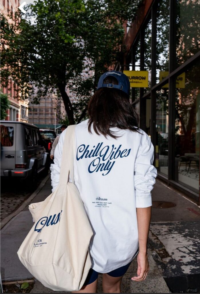 Woman wearing Chill Vibes Only shirt, walking down city street with canvas bag and cap. Urban lifestyle scene.