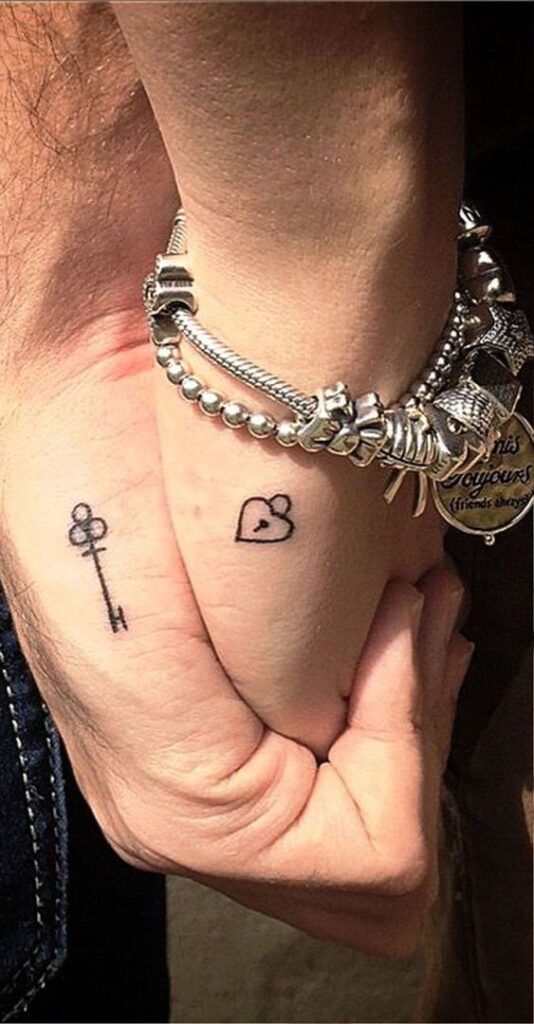 Hands holding with key and heart tattoos, wearing a silver charm bracelet.