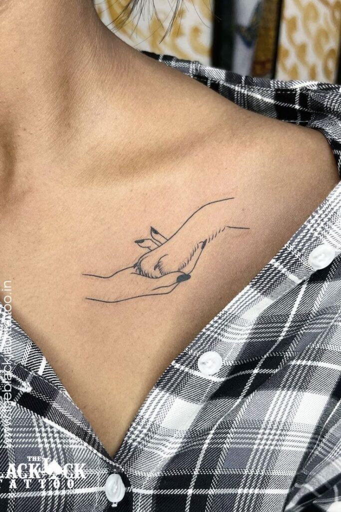 Minimalist tattoo on collarbone of a hand holding a paw, symbolizing friendship and love, on plaid shirt background.