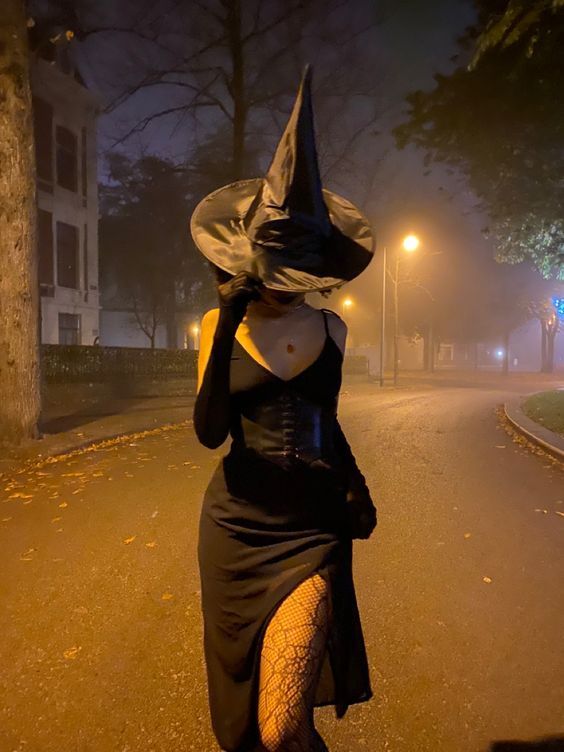 Mysterious witch in a black dress walking down a foggy street at night with streetlights glowing.