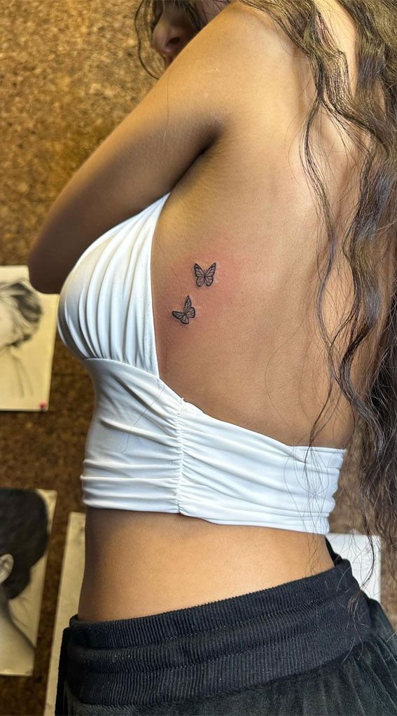 Woman with butterfly tattoos on her back, wearing a white top, showcasing delicate body art in a stylish setting.