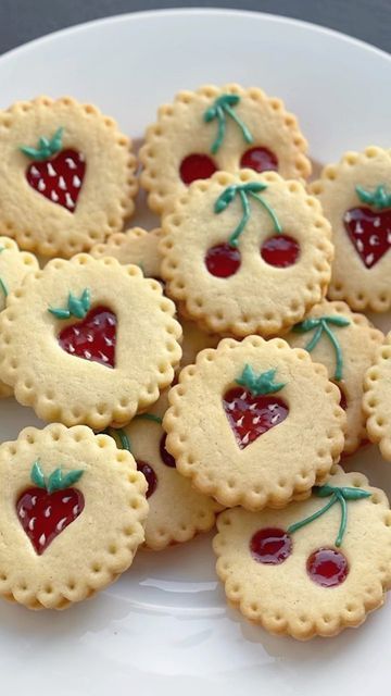 Fruit-shaped cookies with red jam centers featuring strawberries and cherries, arranged on a white plate.