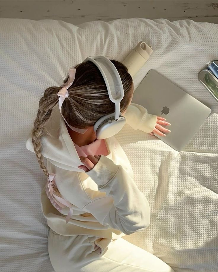 Woman wearing headphones using a laptop in bed, cozy cream outfit with a braided hairstyle and ribbon.