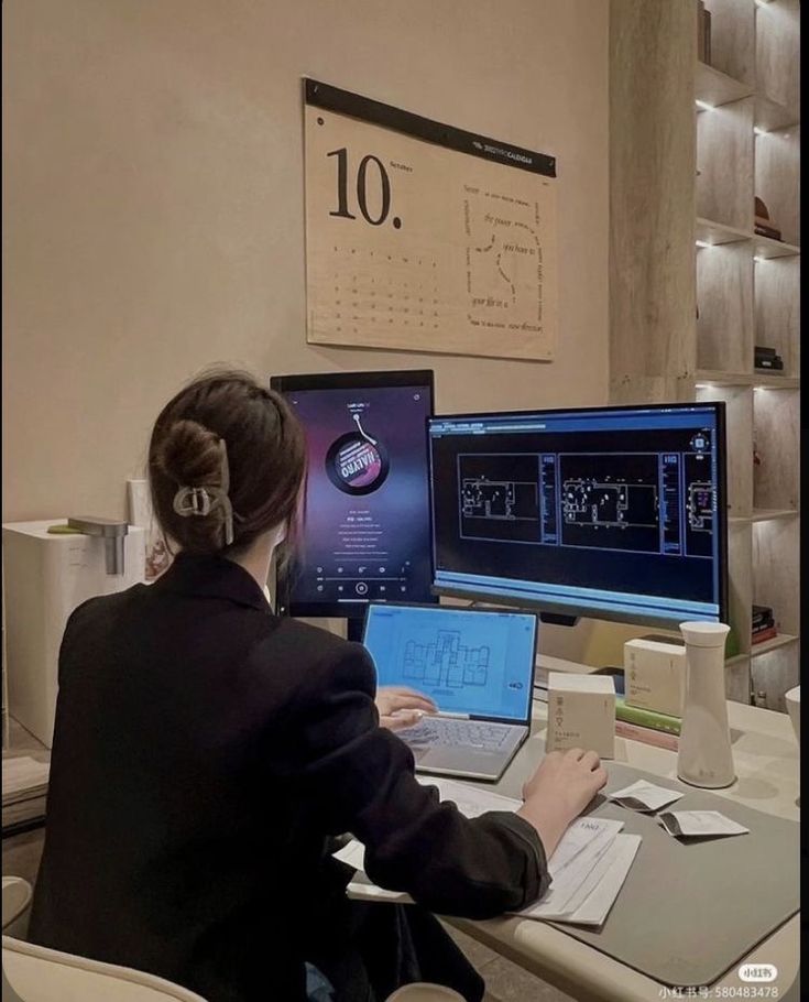 Woman working at desk with multiple screens, architectural plans, and October calendar in office setting.