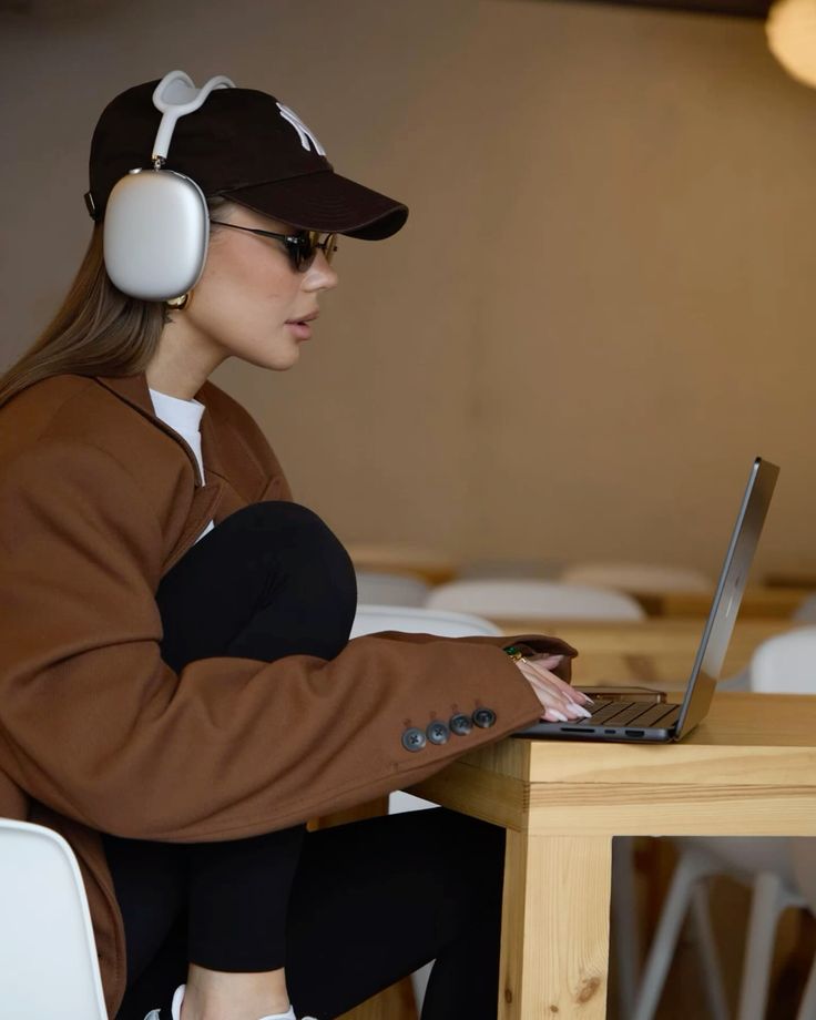 Woman in stylish outfit wearing headphones and working on a laptop at a wooden table, focused and engaged.