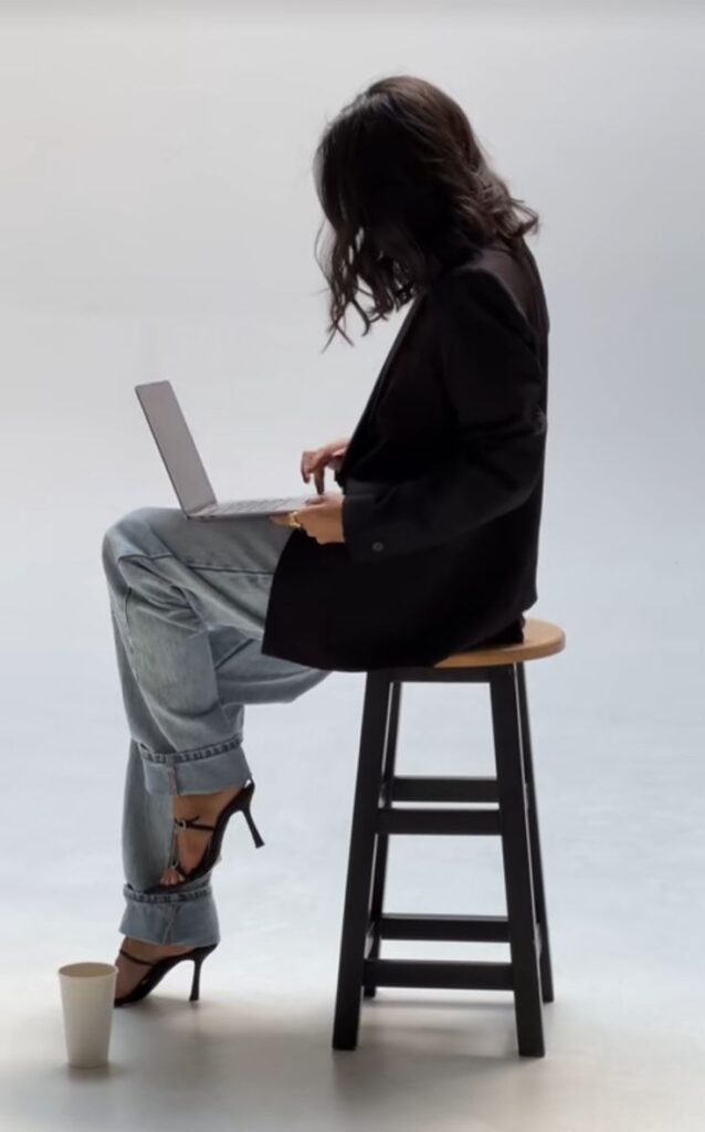 Woman in blazer and jeans typing on laptop while sitting on stool, next to coffee cup, against neutral background.