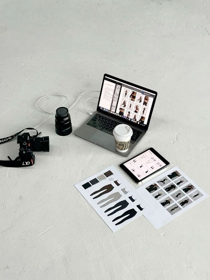 Photography workspace with laptop, camera, coffee cup, tablet, and fashion design printouts on a white surface.