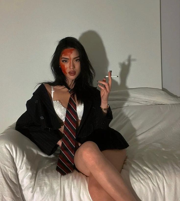 Woman in suit jacket, tie, and lingerie sitting confidently on a bed, holding a cigarette, with bold face paint.