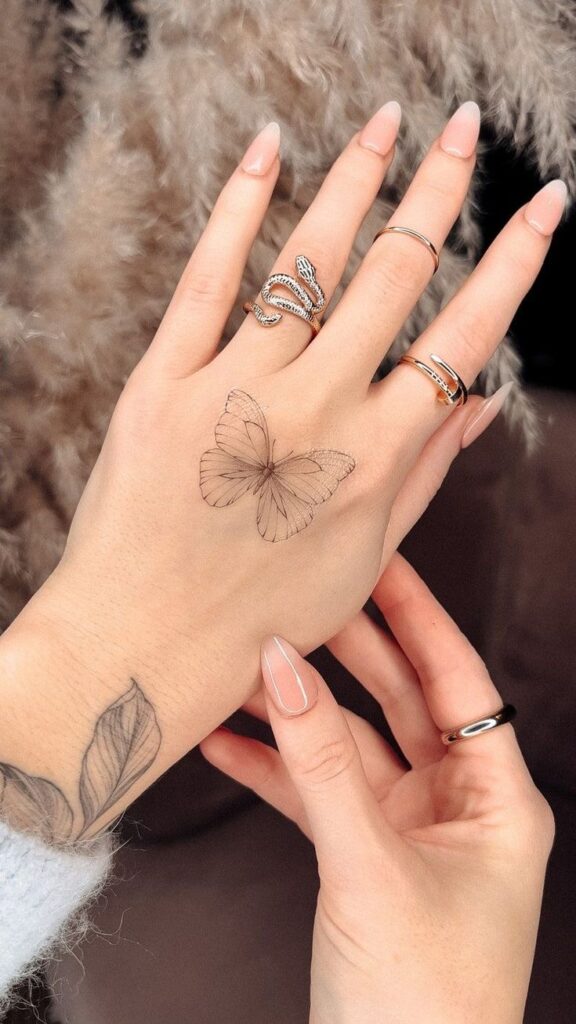 Hand with delicate butterfly tattoo and rings, showcasing detailed nail art against a soft, blurred background.