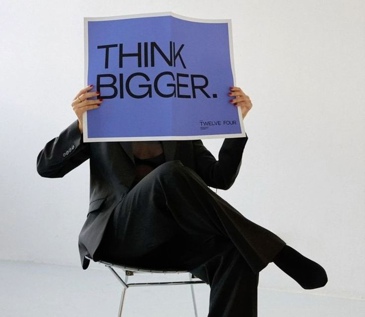 Person in suit holding sign reading THINK BIGGER while sitting on chair; concept of ambition and innovation.