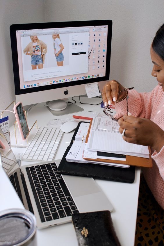 Woman working at desk with laptop, notepad, and screen showing online shopping for fashion items.