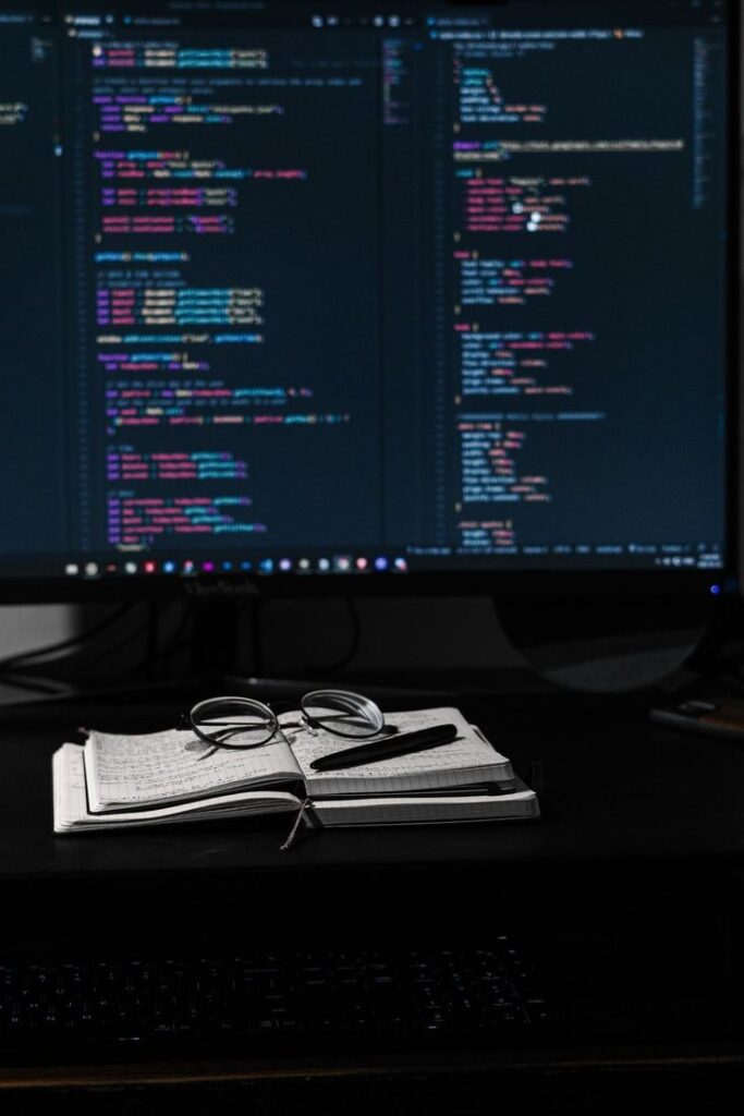 Dual monitors display code; glasses and notebook on desk suggest programming in progress.