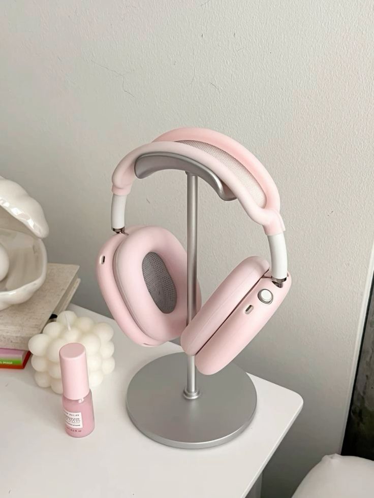 Pink wireless headphones on a stand next to a decorative candle and nail polish on a white desk.