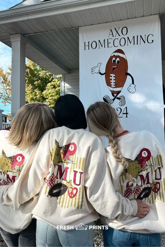 Three people wearing jackets with CMU designs, standing in front of an AXO homecoming banner featuring a football.