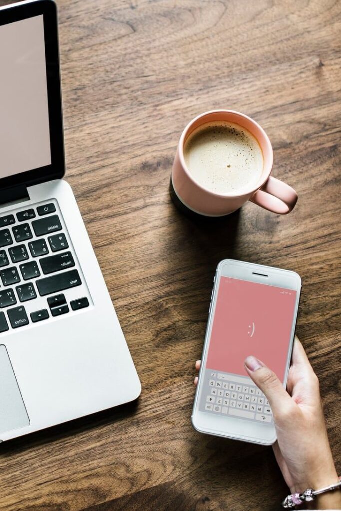 Laptop, smartphone with smiley face screen, and coffee on wooden desk for a cozy, productive workspace vibe.