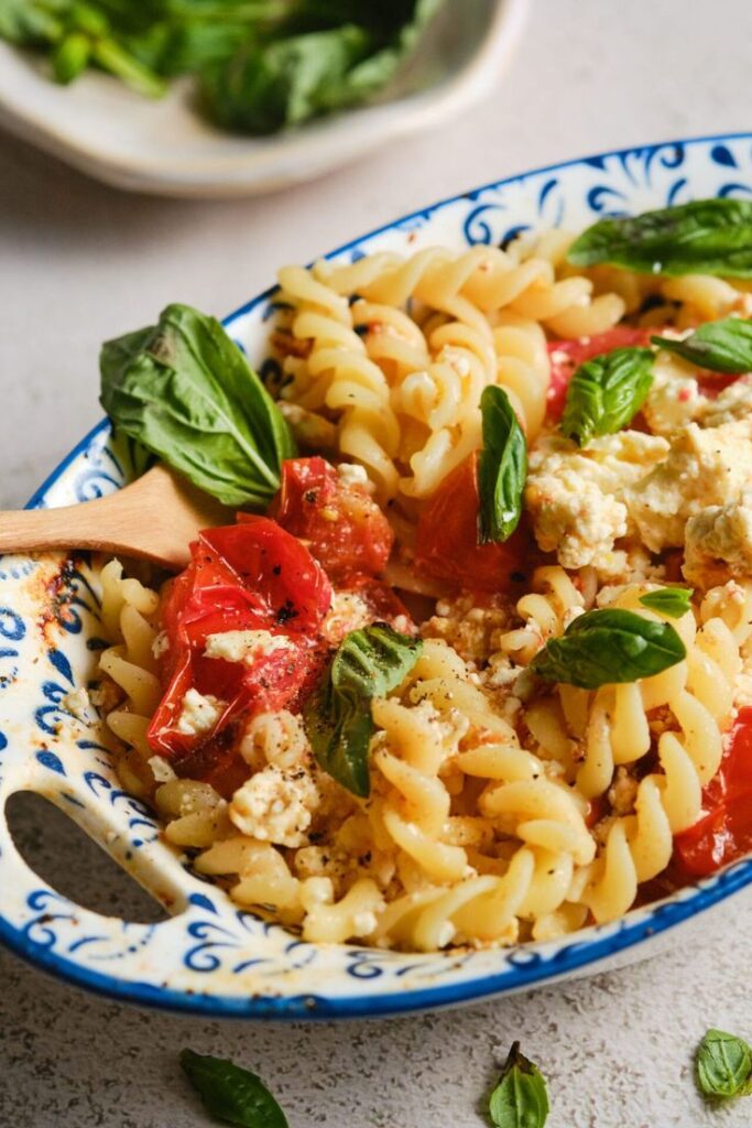 Baked feta pasta with cherry tomatoes, basil, and fusilli in a decorative dish on a light background.