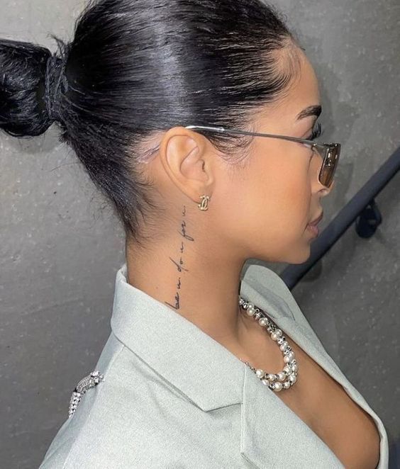 Profile of a stylish woman with sunglasses, elegant necklace, and neck tattoo, wearing a blazer and hair in a bun.