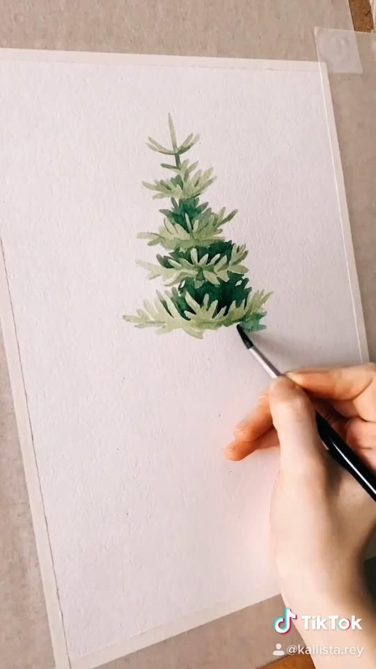 Artist painting a watercolor pine tree on white paper with delicate brush strokes.