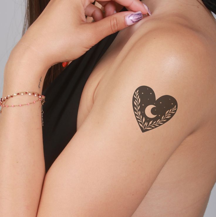 Woman's shoulder tattoo: heart with crescent moon and leaves on upper arm. Black and gold design.