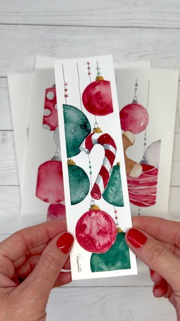 Hands holding a festive bookmark with red and green watercolor ornaments and a candy cane on a white background.