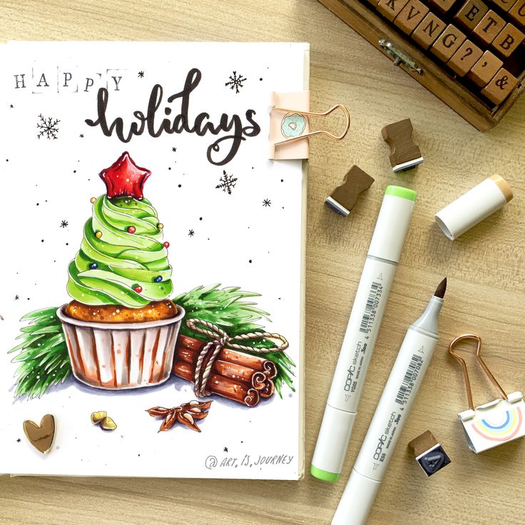 Festive holiday card with cupcake illustration, cinnamon sticks, and art supplies on a wooden desk.