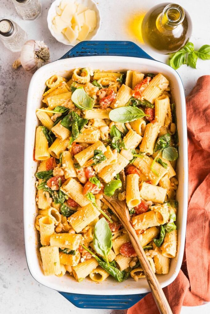 Baked rigatoni pasta with spinach, cherry tomatoes, and cheese in a casserole dish, garnished with fresh basil leaves.