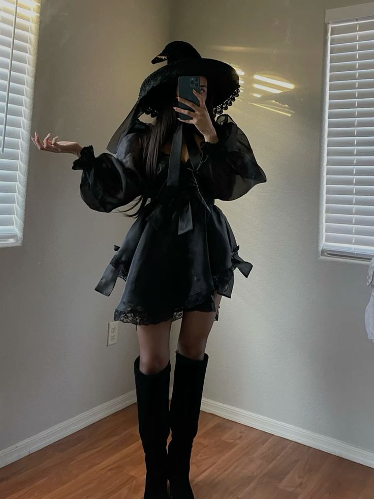 Person in a stylish witch costume with a black hat and boots, standing in a room with wooden floors and bright light.