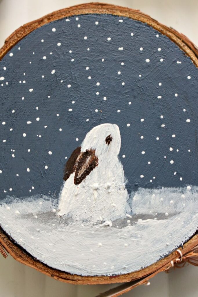 DIY Painted Birch Slice Ornaments Inspired by Maud Lewis - Dans le Lakehouse копия Painting of a snow-covered rabbit on wood with a dark night sky and falling snowflakes. | Sky Rye Design Painting of a snow-covered rabbit on wood with a dark night sky and falling snowflakes.