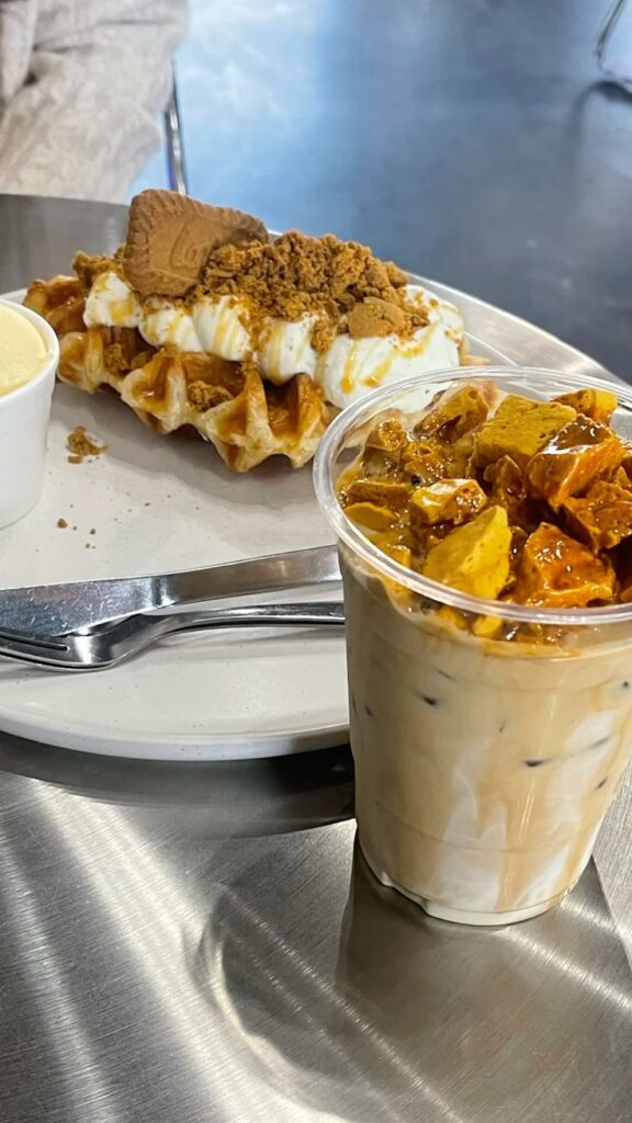 Waffles topped with cream and crumble, iced coffee with honeycomb on a stainless steel table.