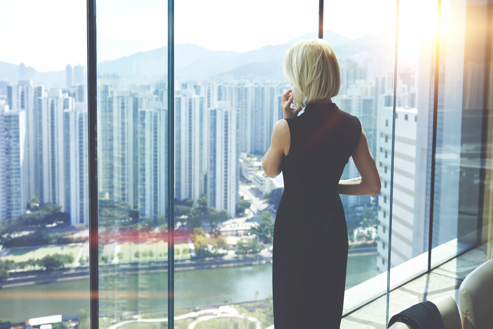 Woman in a sleek black dress gazing at city skyline through large glass window with sunlight streaming in.