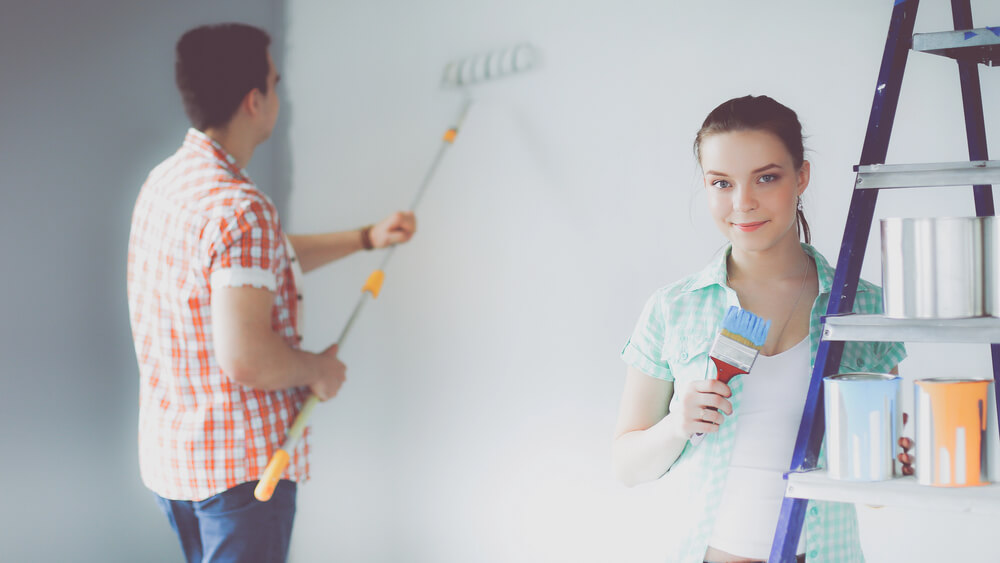 Two people painting a room; woman with brush by ladder, man rolling paint on wall.