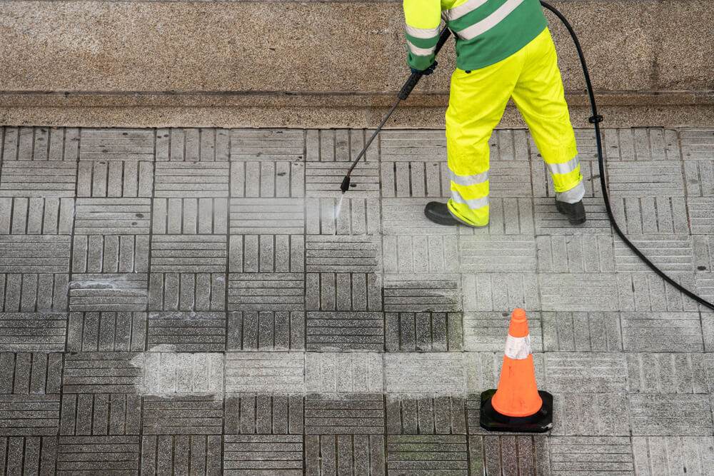 Worker in high-visibility gear pressure washing a sidewalk near a caution cone.