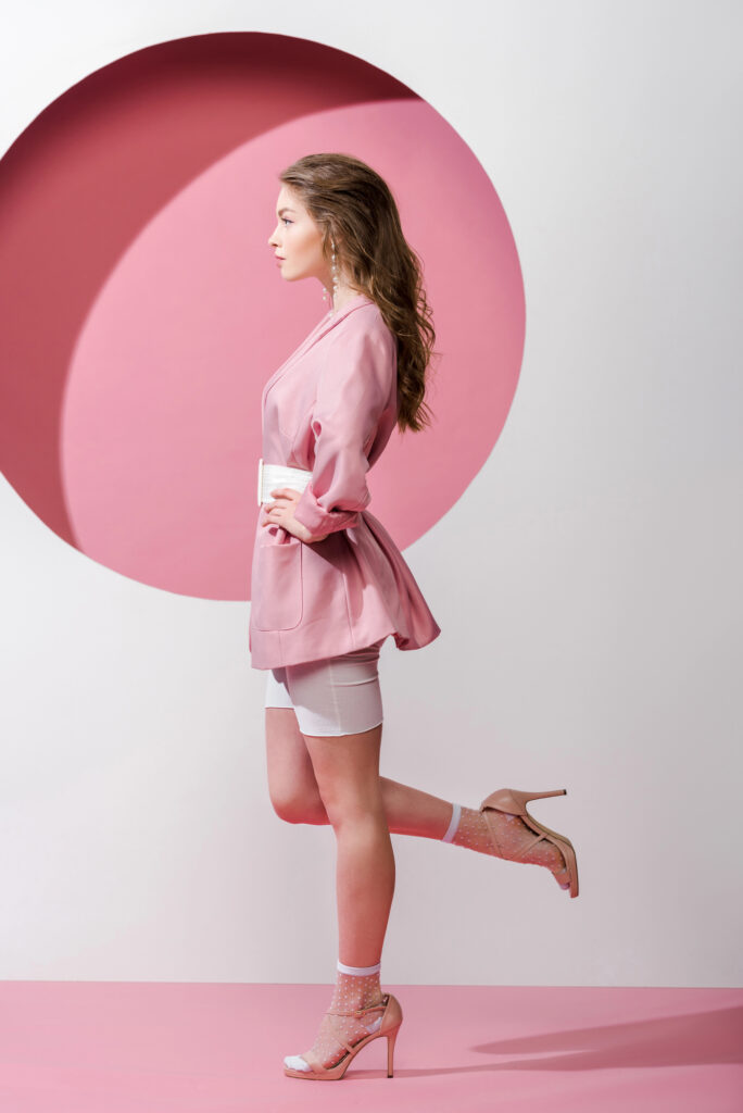 Fashionable woman in pink blazer and heels posing in front of circular pink and white backdrop.