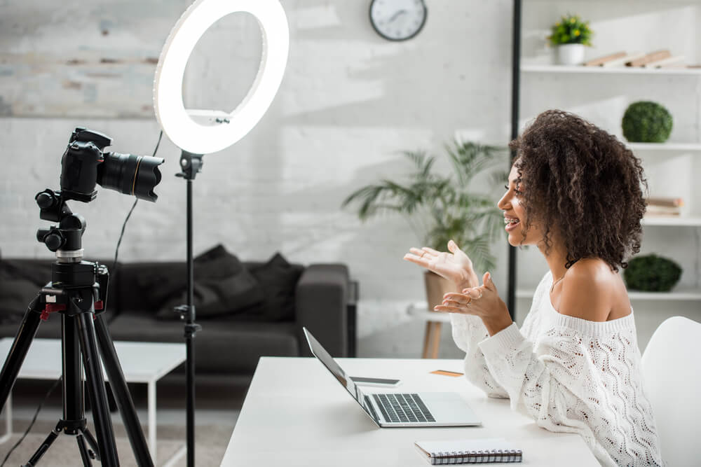 Woman vlogging at home with camera and ring light, sitting at a table with a laptop, creating content for her audience.