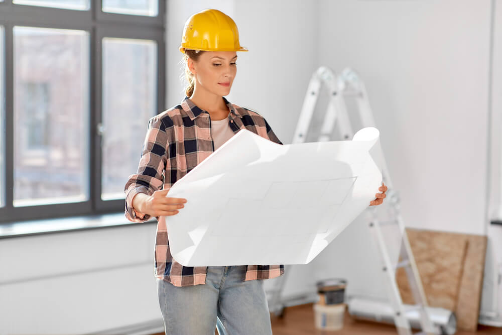 Female construction worker with hard hat examines blueprints in a bright room, ladder and tools in background.