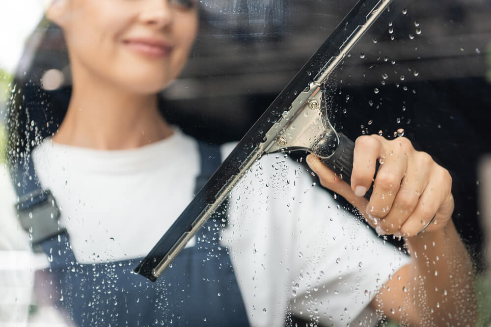 Woman cleaning glass with a squeegee, wearing overalls and smiling, emphasizing efficient window cleaning.