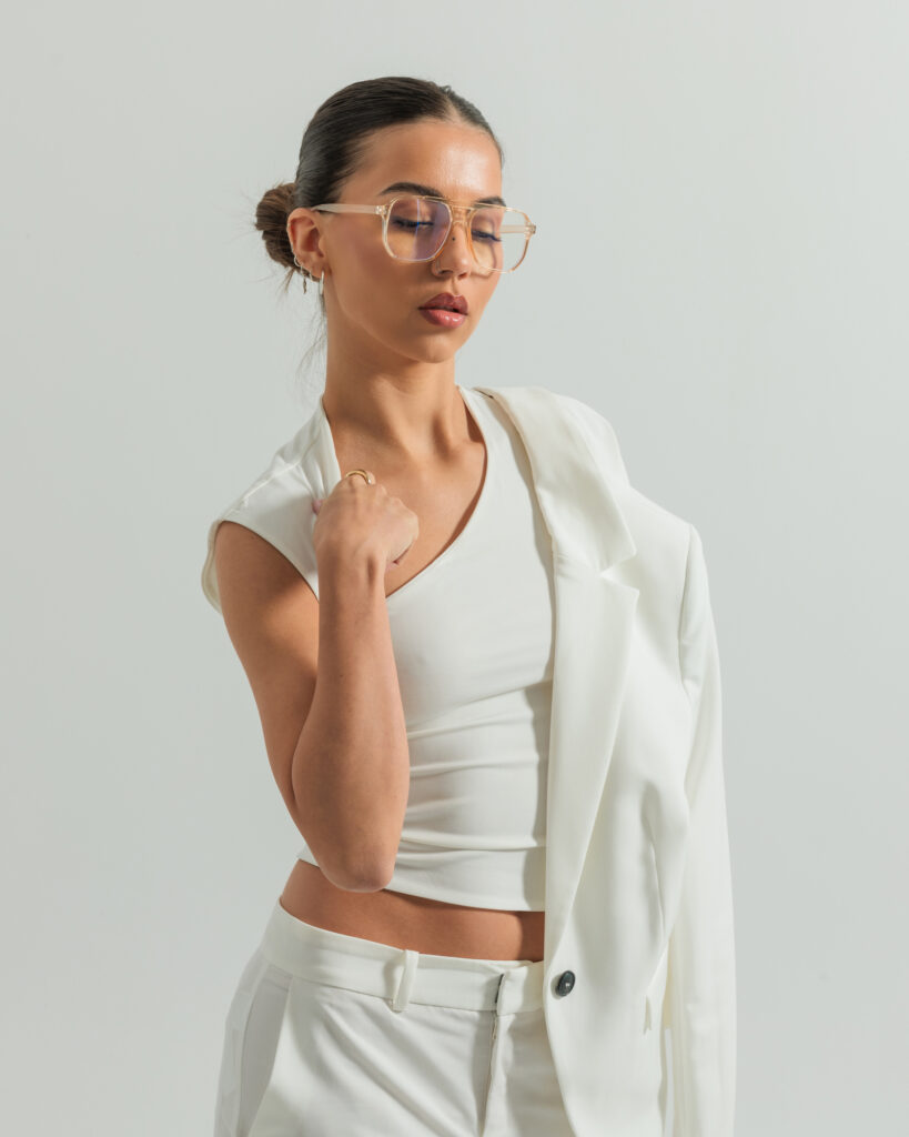 Fashionable woman in white outfit and glasses posing confidently against a neutral background.