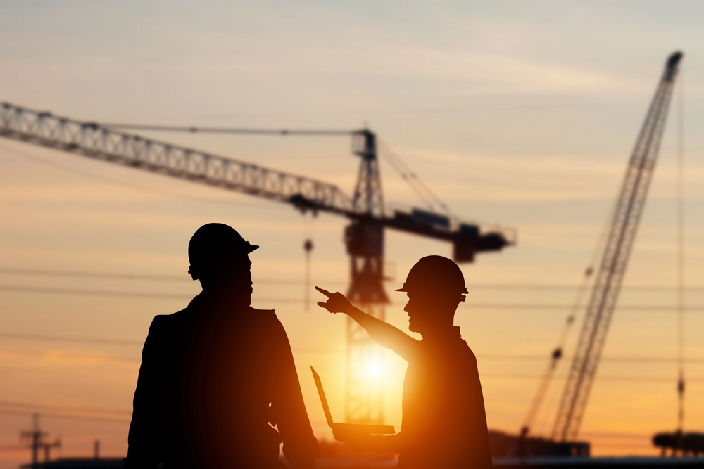 Silhouetted construction workers discussing project at sunset with cranes in the background.