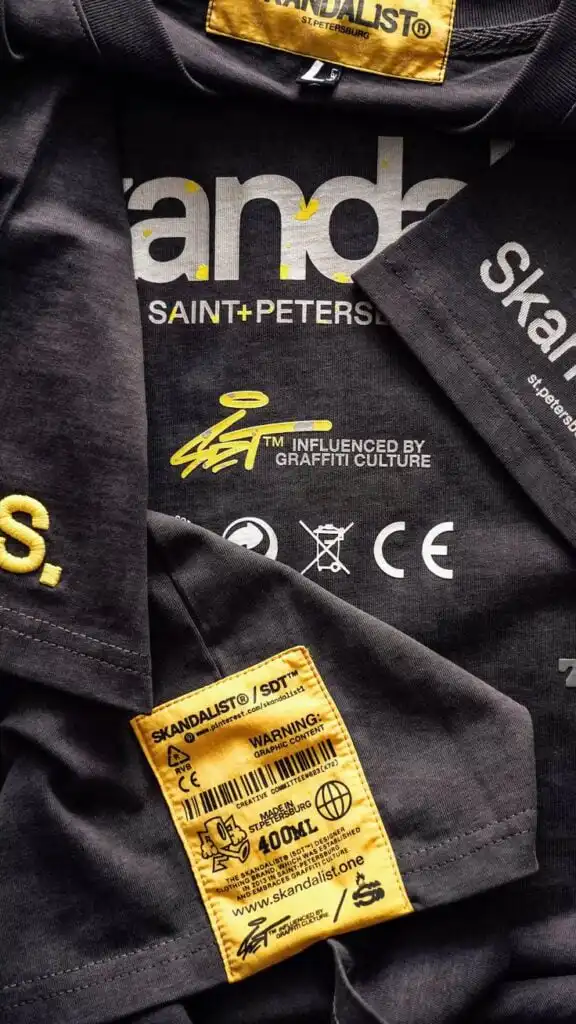 Stacked black Skandalist graphic t-shirts close-up with white SAINT PETERSBURG print, graffiti-style logo and a prominent yellow branded care/warning label