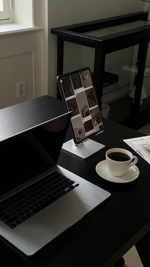 Workspace with a laptop, tablet showing a grid, and a cup of coffee on a black desk by the window.