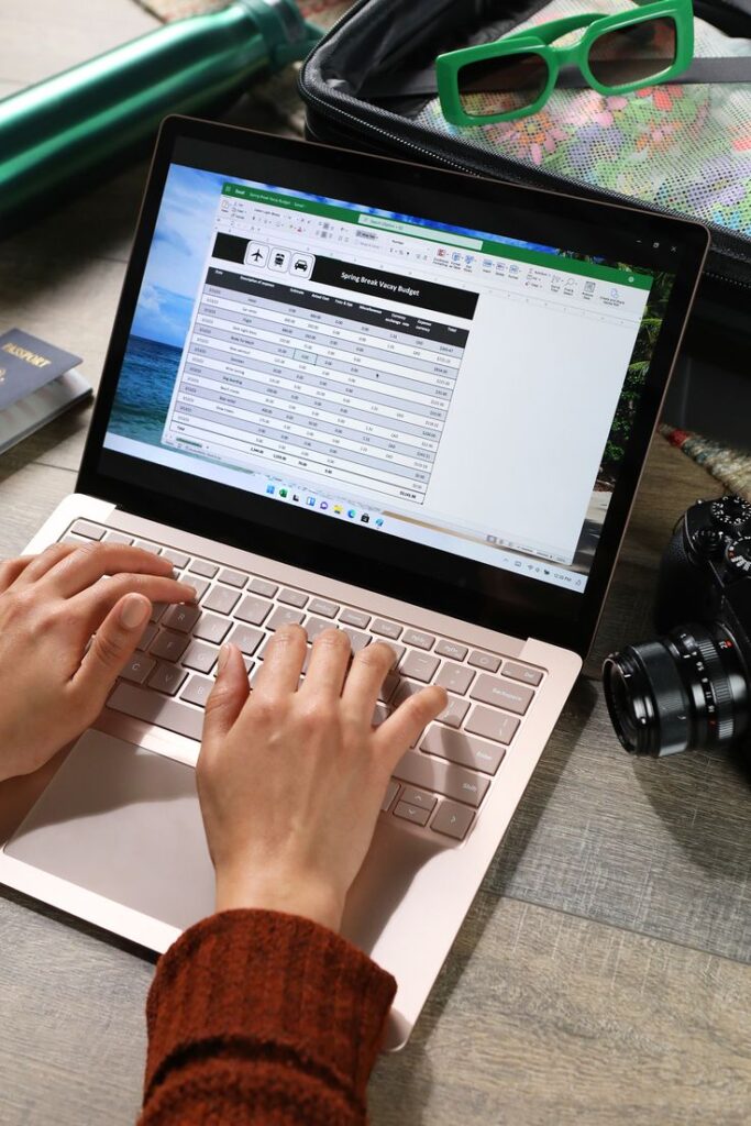 Person using a laptop to manage a vacation budget spreadsheet, surrounded by travel items and camera.